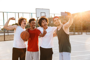Strong multiethnic men smiling and taking selfie on smartphone, while playing basketball at playground outdoor during summer sunny day